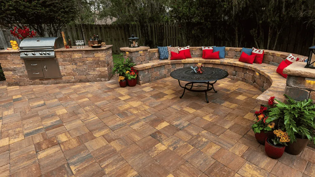 Finished backyard with a spacious brown and yellow paver patio, a stone outdoor kitchen, and a curved seating wall with cushions and a fire pit table.