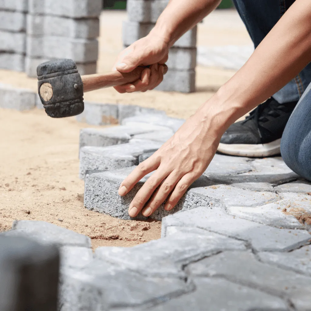 Close-up of a contractor's hands using a rubber mallet to gently tap an interlocking gray paver into the sand base during the paver installation process. Represents professional hardscaping near Miami.