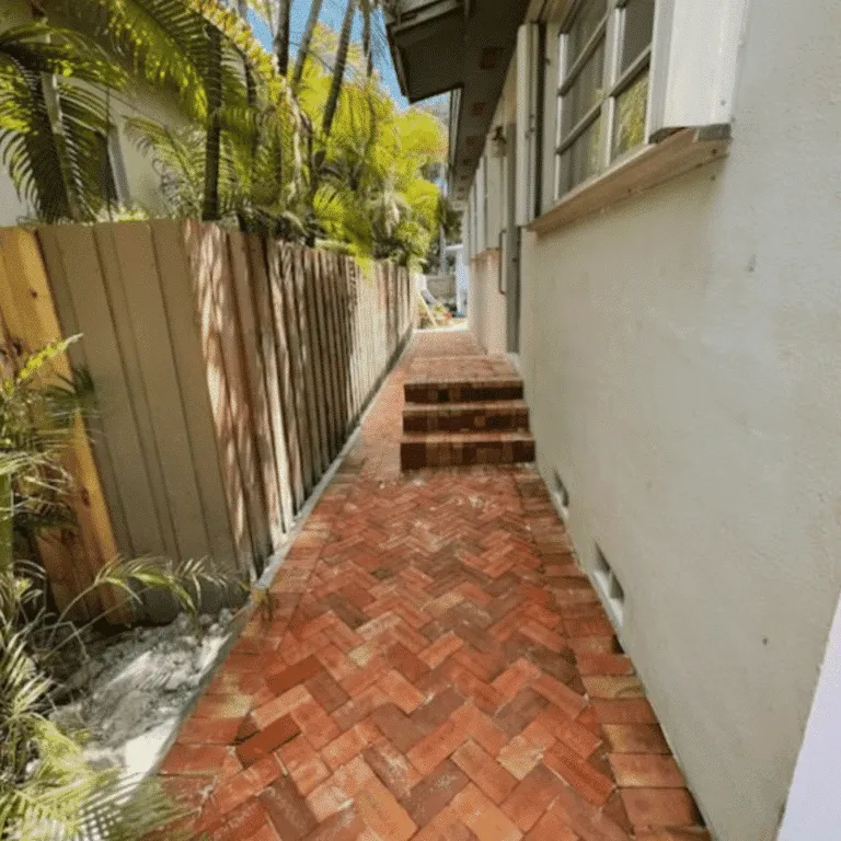 Long, narrow outdoor paver path made of red brick with steps, beside a white house wall and a wooden fence in a tropical area.
