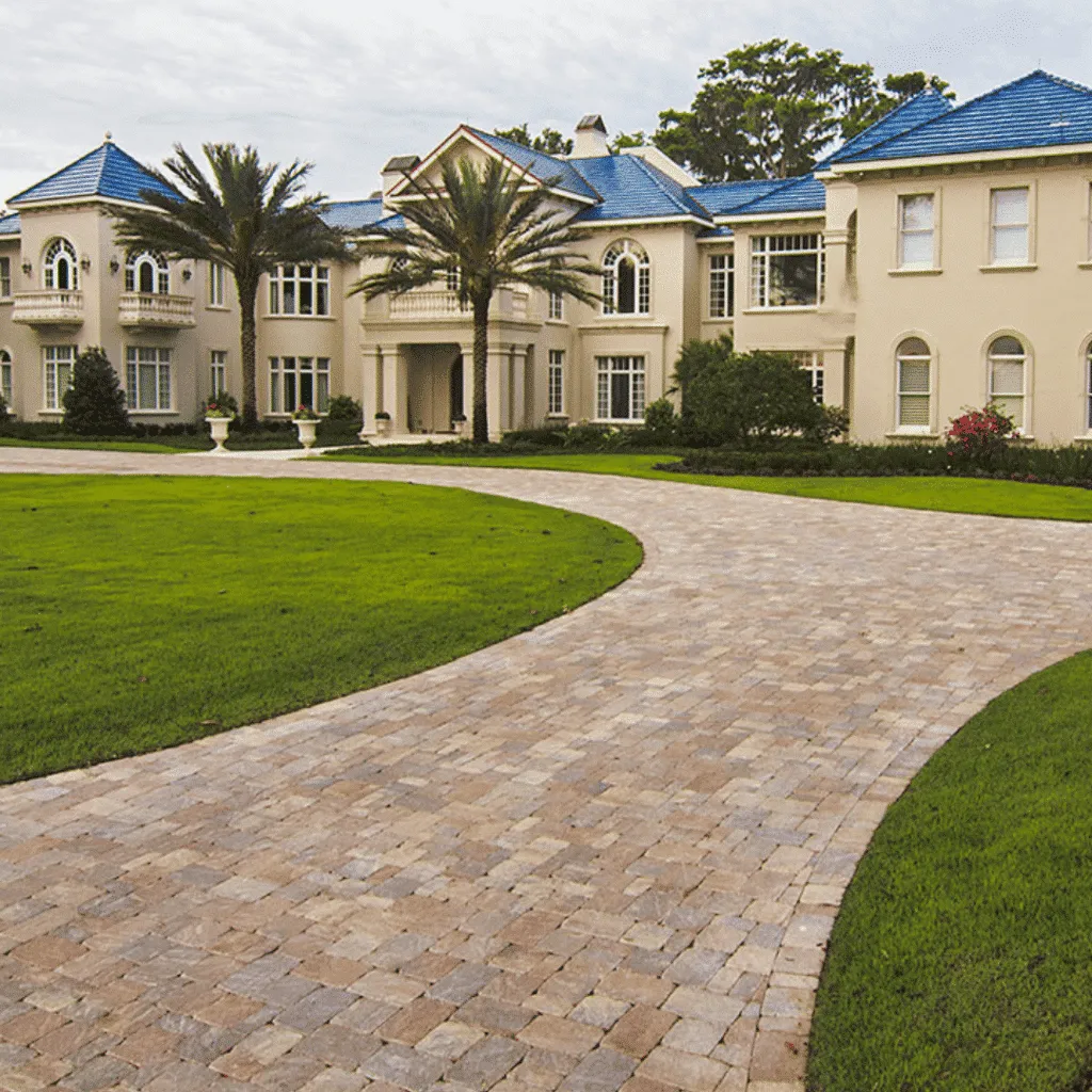 Long, curved paver driveway leading to a mansion with stucco walls and blue roofs, surrounded by palm trees