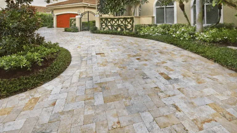 Curved residential driveway made of large, rectangular, light-colored, multi-tone travertine pavers set in a staggered or modular pattern. The driveway leads to a Mediterranean-style home with an orange garage door and lush landscaping. Example of premium paver and tile installation in Miami.