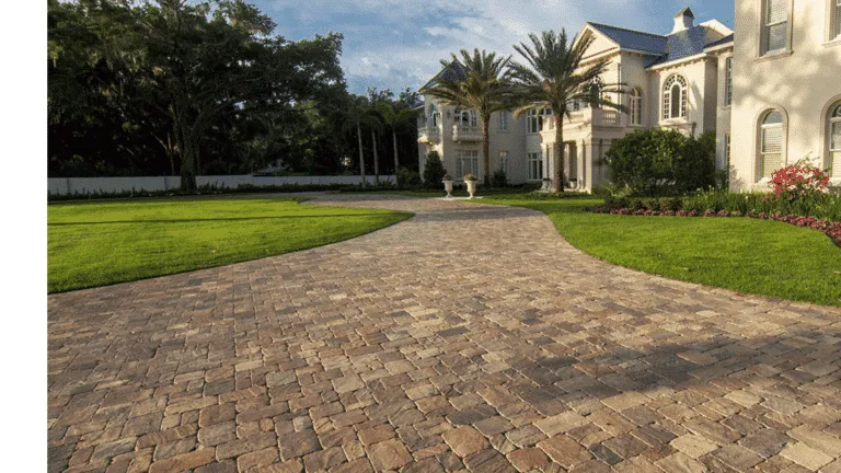 Large, curving residential driveway made of rough-textured, multi-colored tan and brown pavers. The driveway is wide and leads to a large, luxurious, light-colored home surrounded by a manicured green lawn and palm trees. Example of high-end paver installation in Miami, Florida.