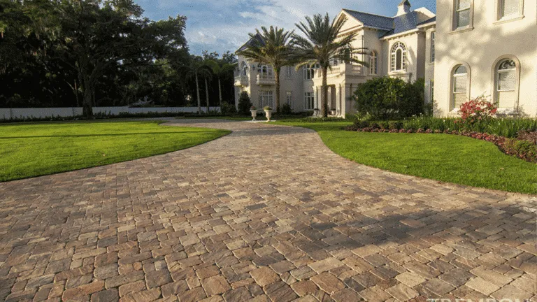Wide, curving residential driveway made of textured, multi-color brown and tan pavers. The driveway is surrounded by lush, manicured lawn and leads to a large, light-colored luxury home.