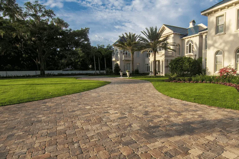 Wide, curving residential driveway made of textured, multi-color brown and tan pavers. The driveway is surrounded by lush, manicured lawn and leads to a large, light-colored luxury home. Example of high-quality paver installation in Miami, Florida.