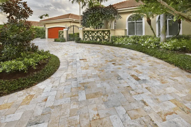 Curved residential driveway made of light, multicolored, rectangular natural stone pavers (possibly travertine) laid in a herringbone pattern. The driveway leads to a Mediterranean-style home with lush landscaping.