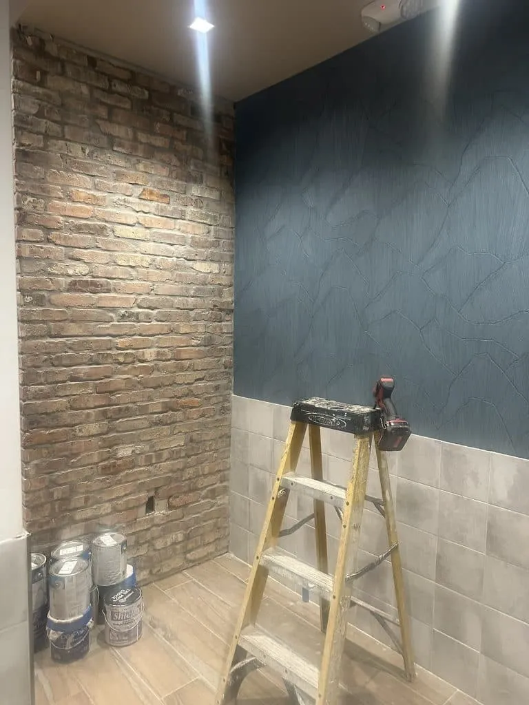 Interior room under construction showing an exposed brick wall, a blue textured wall, and light wood-look floor tiles.