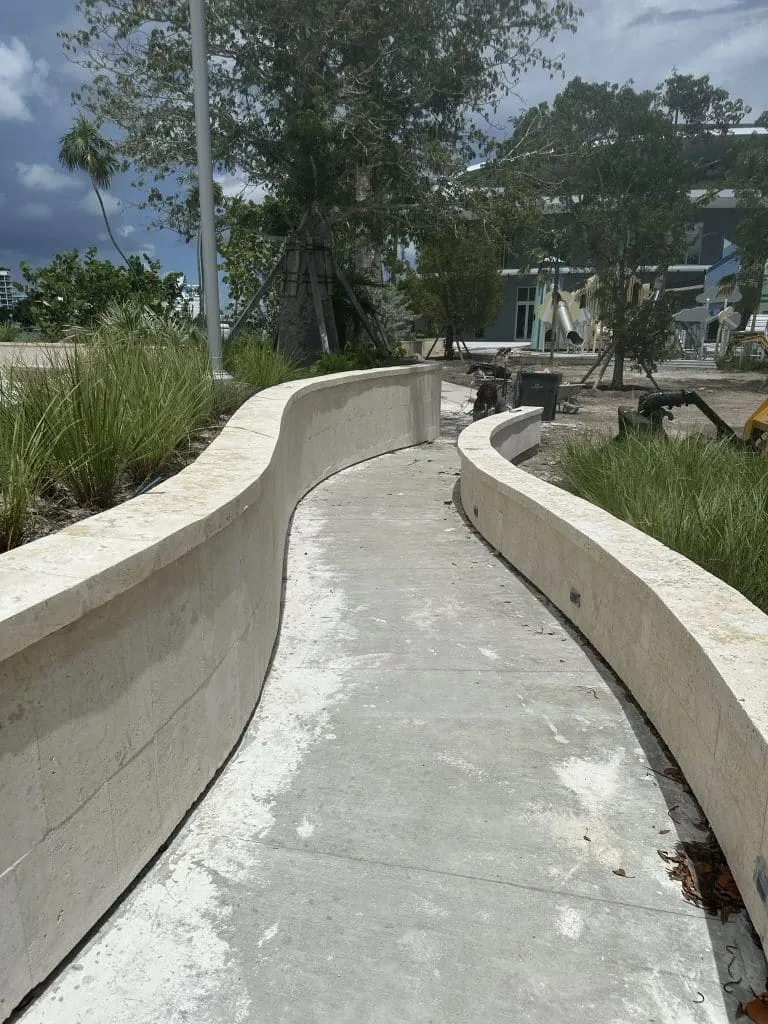 Curved concrete walkway bordered by two low, light-colored limestone retaining walls in a landscaped park.