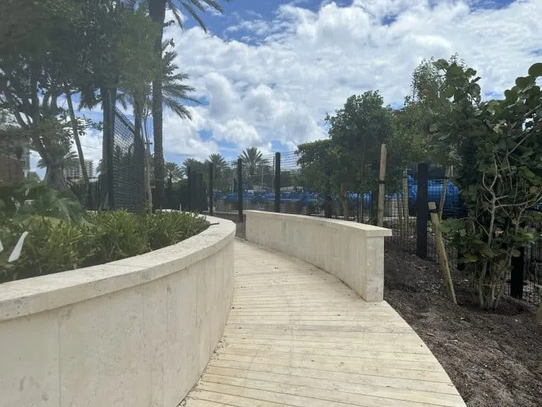 Curved elevated wooden boardwalk pathway bordered by low, light-colored stone walls under a sunny, cloudy sky with palm trees.
