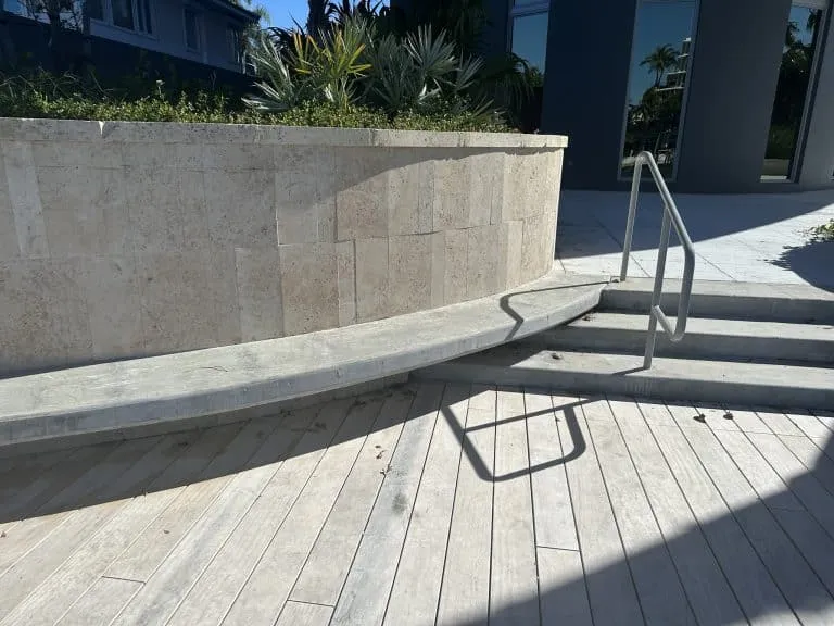 Curved stone tile planter wall and concrete bench next to light wood-look plank pavers and concrete steps with a metal railing.