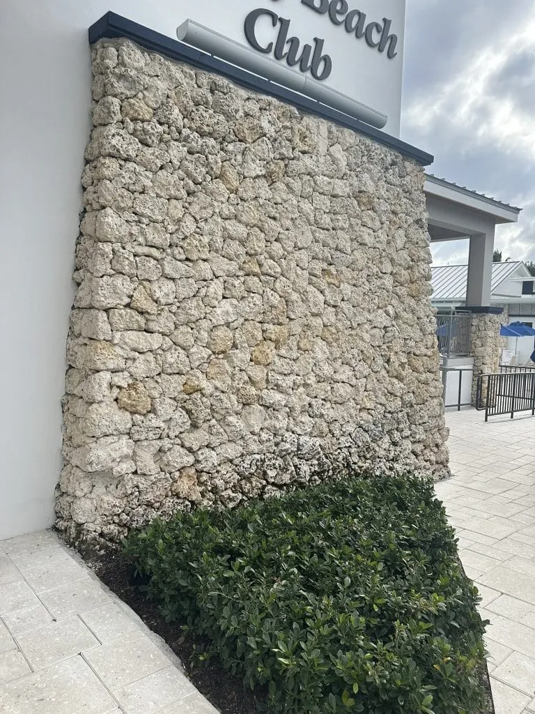 Large exterior wall covered in rough, light-colored natural stone veneer next to a clean, light gray paver walkway at a beach club.