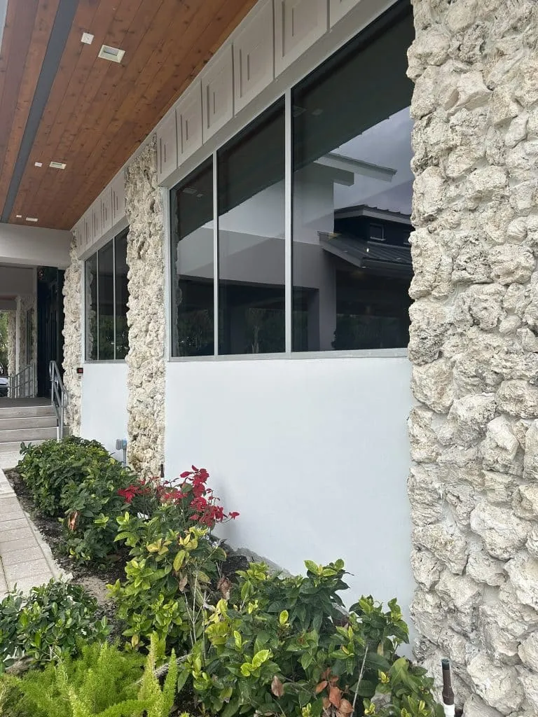 Building exterior with columns clad in rough natural coral stone veneer, a wood ceiling, and a light-colored paver walkway.